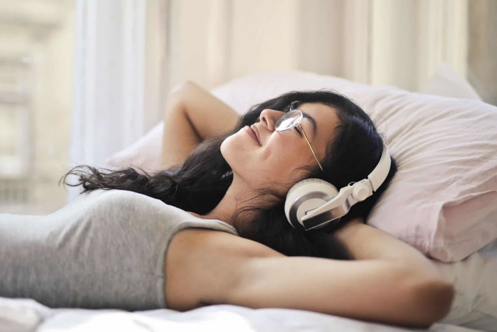 Kobieta leży na łóżku. Jest zrelaksowana i słucha muzyki w słuchawkach nausznych.